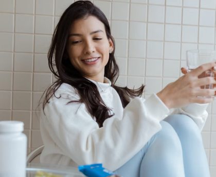 Smiling woman holding a glass while seated, with pill bottles on the table.