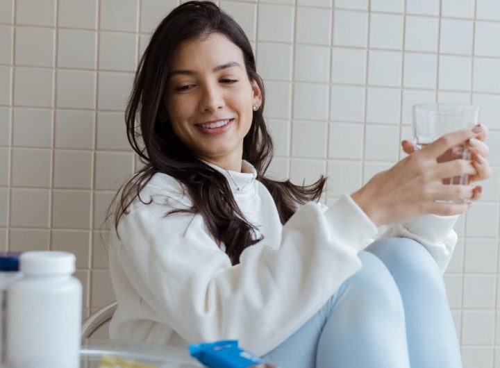 Smiling woman holding a glass while seated, with pill bottles on the table.