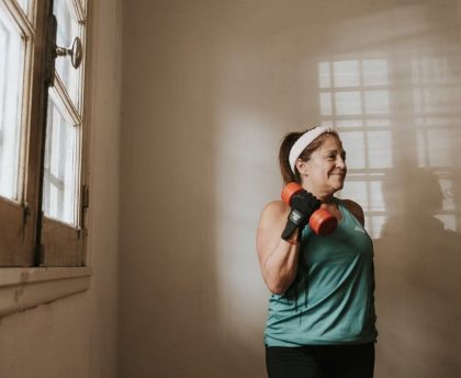 Senior woman in a brightly lit room exercises with dumbbells, promoting fitness at home.