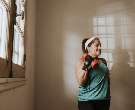 Senior woman in a brightly lit room exercises with dumbbells, promoting fitness at home.
