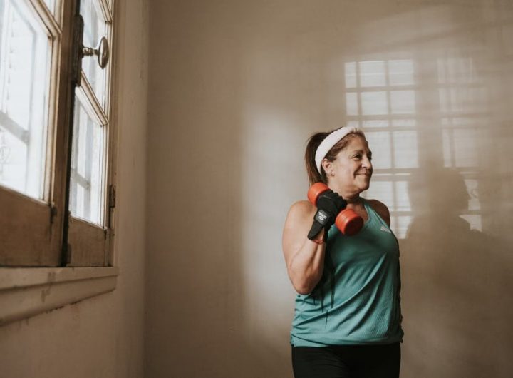 Senior woman in a brightly lit room exercises with dumbbells, promoting fitness at home.